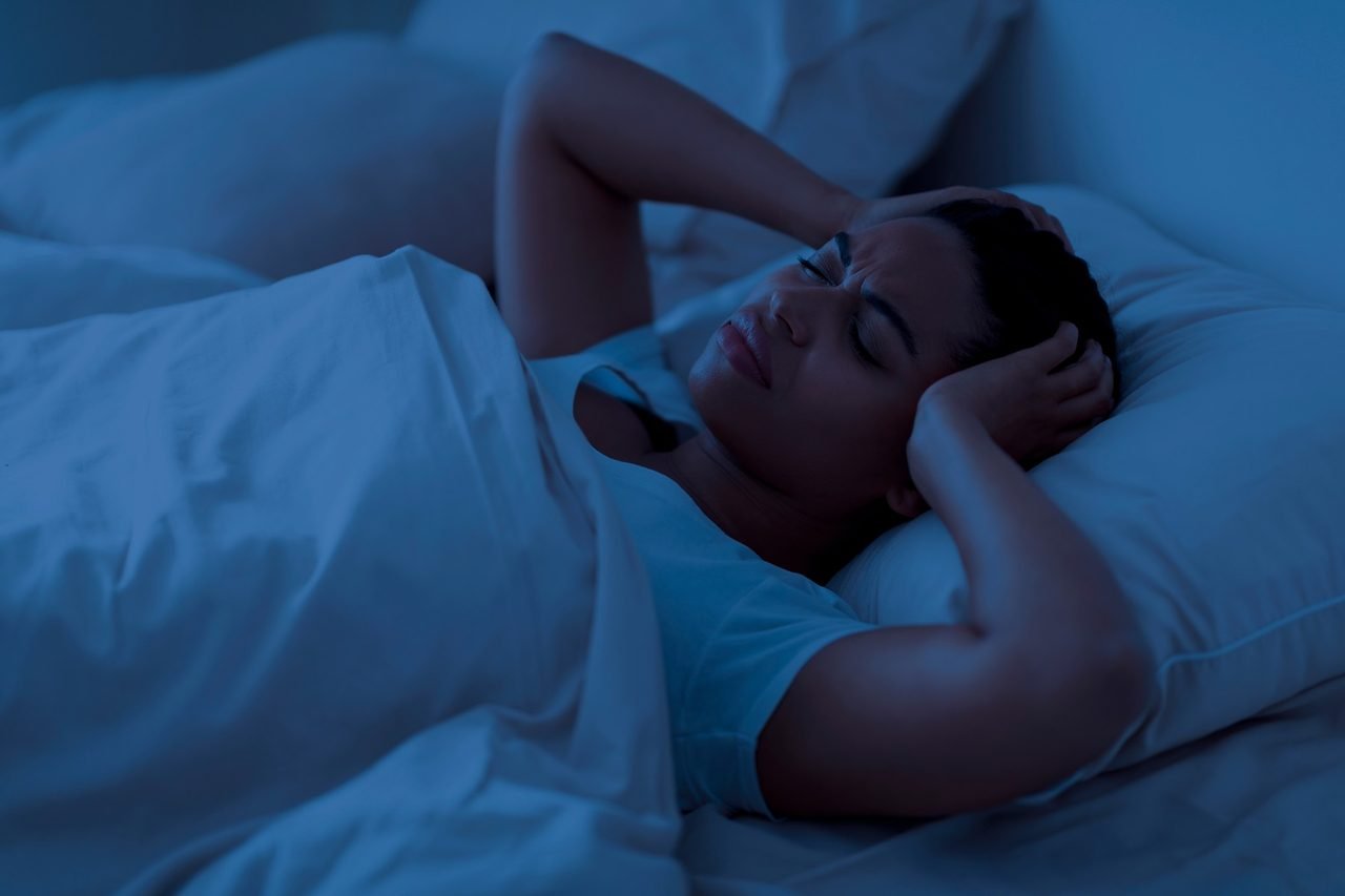 Woman lying awake in bed at night pressing her hands against her head in discomfort